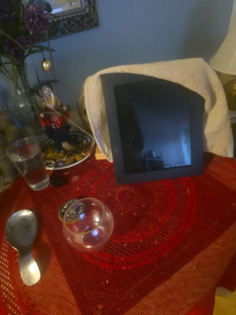 A black framed scrying mirror draped with a light-colored cloth rests on a red patterned altar cloth alongside a clear glass bowl, a metal spoon, a glass of water, and a figurine with flowers for a divination setup.