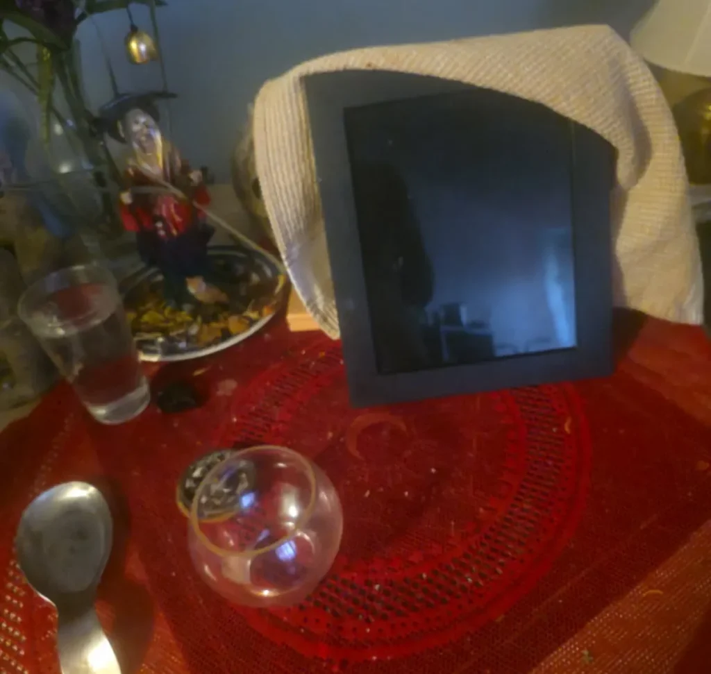 A black framed scrying mirror draped with a light-colored cloth rests on a red patterned altar cloth alongside a clear glass bowl, a metal spoon, a glass of water, and a figurine with flowers for a divination setup.
